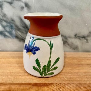 May include: A small, handcrafted ceramic vase with a white body and a dark red rim. The vase features a hand-painted blue flower with green leaves and stem. The vase is sitting on a wooden surface.