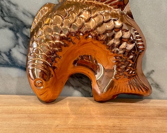 Op de afbeelding: Een glanzende koperkleurige visvormige mal met gedetailleerde reliëfschubben, vinnen en een rond oog. De mal is voorzien van een klein metalen lusje aan de bovenkant om op te hangen en heeft een reflecterend metalen oppervlak met een gebogen vorm.