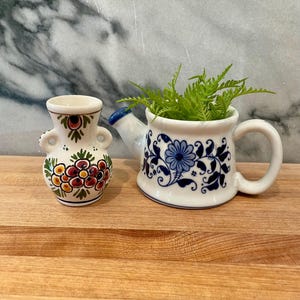 May include: Two ceramic decorative pieces sit on a wooden surface. A small vase with a floral design in red, yellow, and green sits next to a white watering can-shaped planter with blue floral patterns. The planter holds green fern.