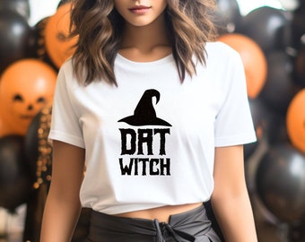 Può includere: Un top bianco corto con un cappello da strega nero e la scritta "DAT WITCH" stampata sul davanti. La modella indossa una gonna nera e si trova davanti a un muro decorato con palloncini neri e arancioni.