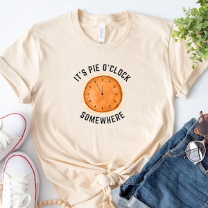 May include: A beige t-shirt with the text "It's Pie O'Clock Somewhere" and a pie clock graphic. The shirt is paired with denim shorts, white sneakers, and a lollipop.
