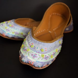 May include: A pair of light blue embroidered flat shoes with colourful floral designs and beaded accents.