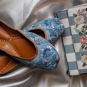 May include: A pair of light blue embroidered flat shoes with brown leather soles. The shoes are decorated with sequins and beads. The shoes are next to a blue and white striped clutch with floral embroidery.
