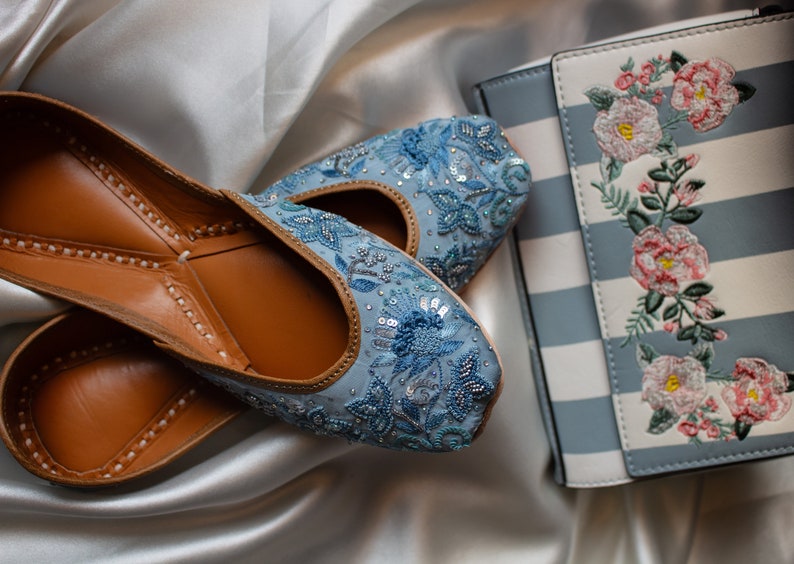 May include: A pair of light blue embroidered flat shoes with brown leather soles. The shoes are decorated with sequins and beads. The shoes are next to a blue and white striped clutch with floral embroidery.