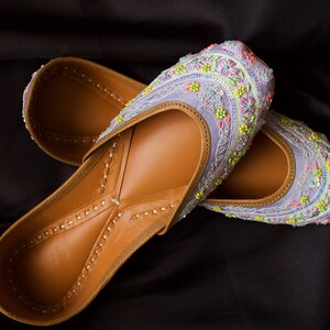 May include: A pair of purple embroidered flat shoes with colourful floral designs. The shoes are made of leather and have a brown interior.