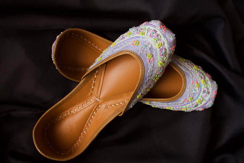 May include: A pair of purple embroidered flat shoes with colourful floral designs. The shoes are made of leather and have a brown interior.