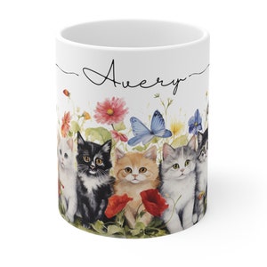 May include: White ceramic mug with a black cursive inscription "Avery" and a colorful illustration of kittens in a field of flowers and a butterfly.