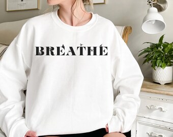 Może przedstawiać: Biała bluza z nadrukiem słowa "BREATHE" w czarnych drukowanych literach. Litery są ozdobione małymi, stylizowanymi obrazami motyli, drzew i innych elementów inspirowanych naturą.
