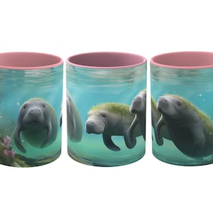 Puede incluir: Una taza de cerámica rosa y blanca con un diseño de tres manatíes nadando bajo el agua. Los manatíes son grises con algas verdes en la espalda. El fondo es azul claro con burbujas.