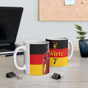 Puede incluir: Taza de cerámica blanca con un diseño de la bandera alemana y un jugador de fútbol con una camiseta roja. La taza muestra el nombre "Wirtz" y el número "7". También se ve un emblema negro y dorado.