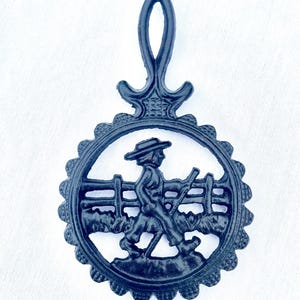 May include: Black cast iron trivet with a handle. The trivet features a circular design with a scalloped edge and a silhouette of a person walking past a fence. The handle has a decorative design.