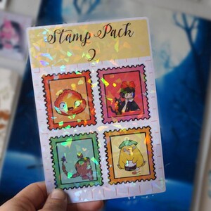 Puede incluir: Una hoja de pegatinas hologr&aacute;ficas con el texto "Stamp Pack 2" y cuatro sellos coloridos con personajes de la pel&iacute;cula animada "El castillo ambulante".