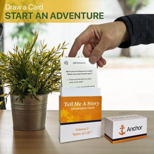 Puede incluir: Un juego de cartas con el texto "Tell Me A Story" y "Start an Adventure". La caja es blanca y naranja, con un diseño de piezas de rompecabezas. Una mano está sacando una carta que dice "Adventures".