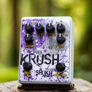 Puede incluir: Pedal de efectos para guitarra plateado con perillas negras y diseño de salpicaduras moradas. El texto "KRUSH" es prominente, con "SHOKST PEDALS" debajo. El pedal tiene múltiples perillas de control.