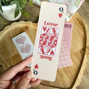 Peut inclure: Un marque-page rose avec un design de carte de jeu de la reine de c&oelig;ur rouge. La carte indique "Book Lover" et a un c&oelig;ur rouge dans le coin.