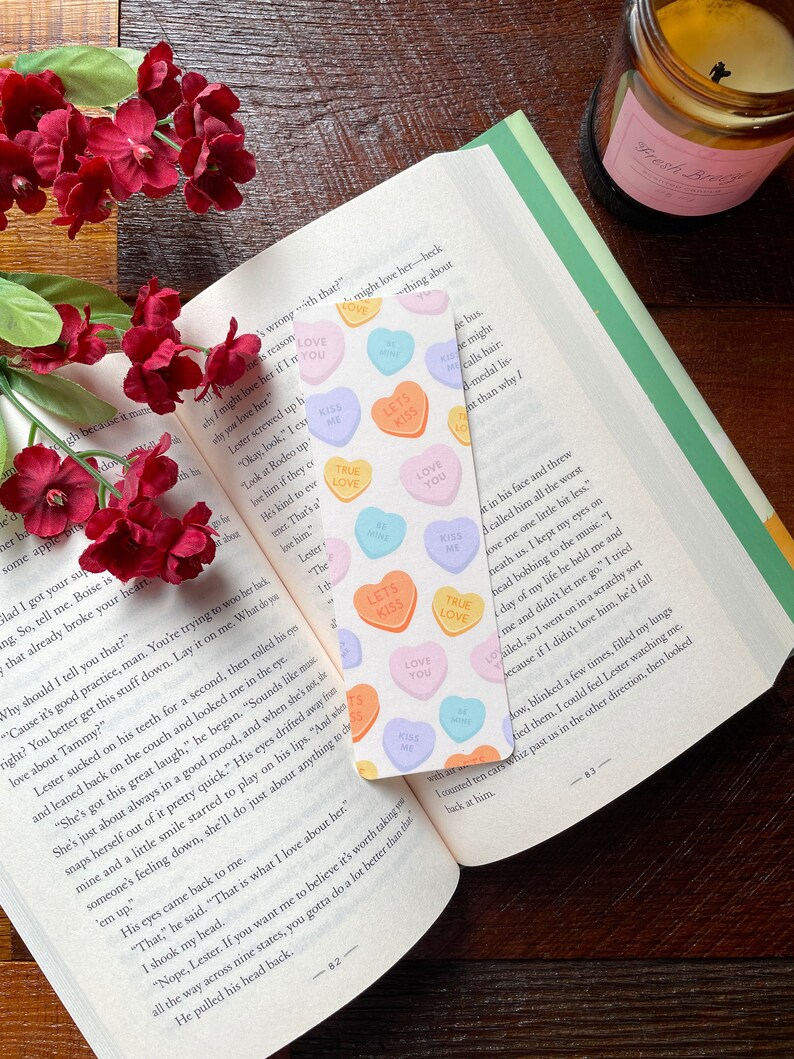 May include: A bookmark with candy heart designs, featuring phrases like "Kiss Me" and "Love You", is placed in an open book. Red flowers and a candle are also visible, creating a romantic scene.