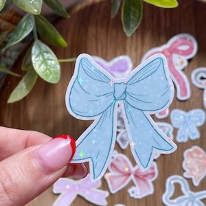 May include: A light blue bow sticker with a holographic glitter effect, held by a hand. Several other bow stickers in various pastel colors are scattered around, including pink, purple, and peach. The stickers are on a wooden surface.