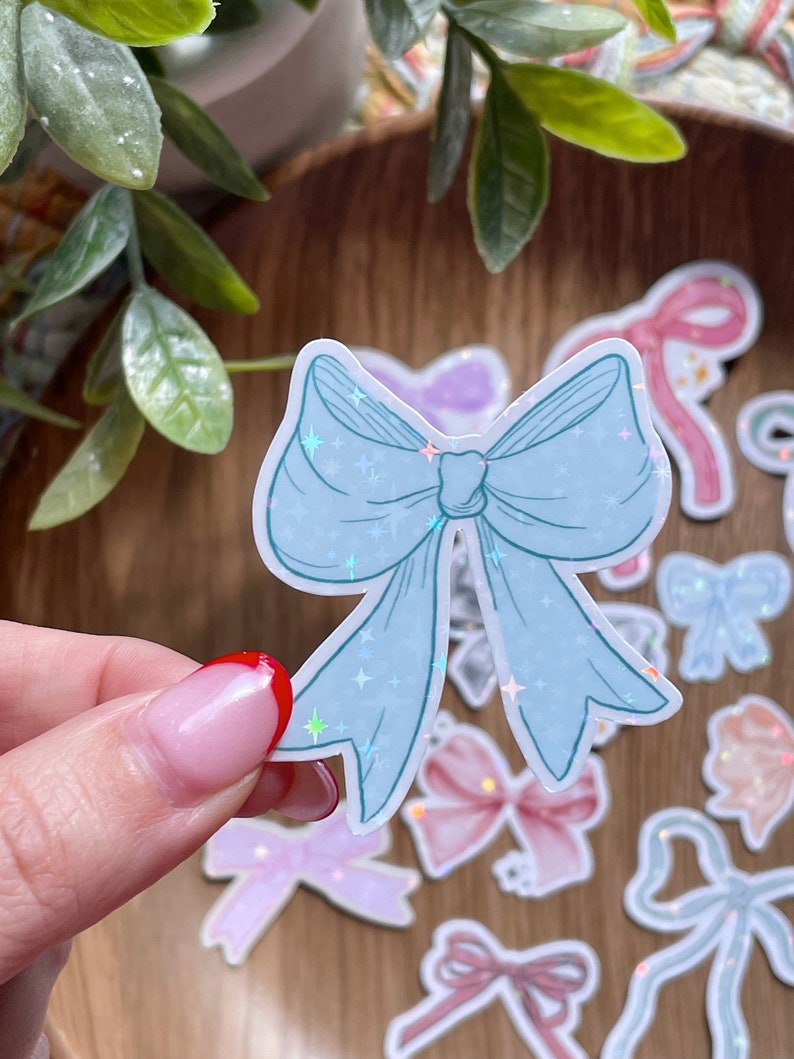 May include: A light blue bow sticker with a holographic glitter effect, held by a hand. Several other bow stickers in various pastel colors are scattered around, including pink, purple, and peach. The stickers are on a wooden surface.