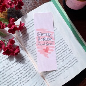 May include: A pink bookmark with the text "Classy Ladies Read Smut" and a pink heart graphic. The bookmark is in an open book with red flowers. The bookmark is a reading accessory.