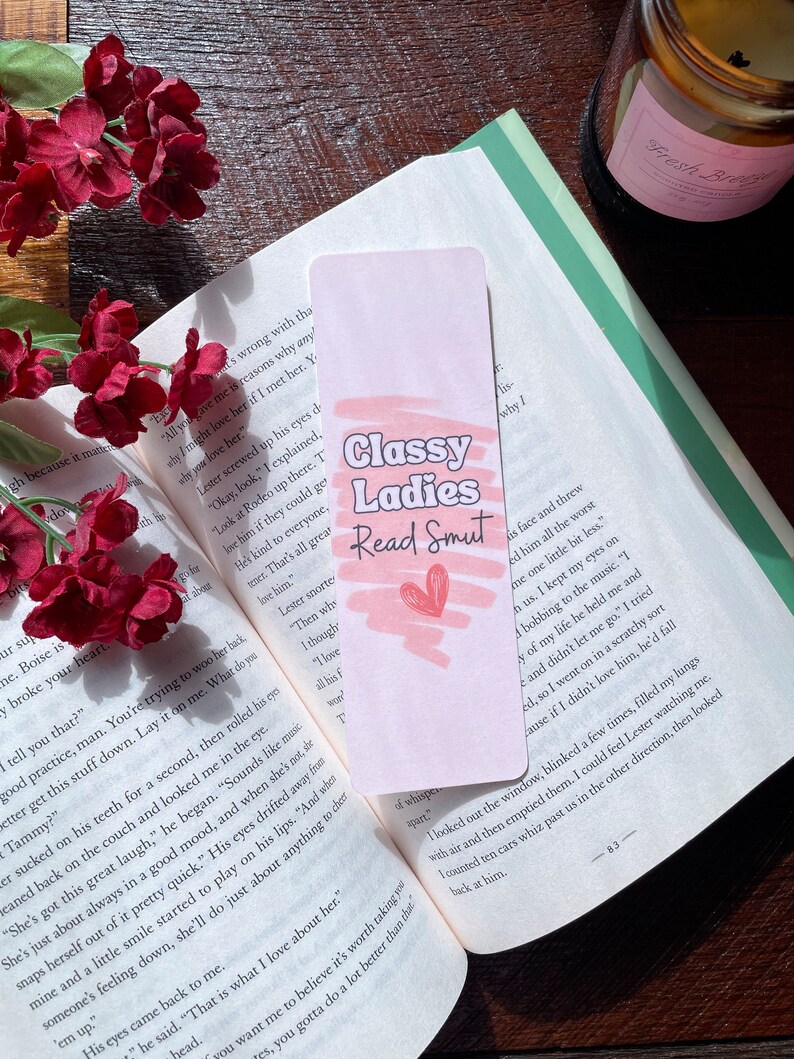 May include: A pink bookmark with the text "Classy Ladies Read Smut" and a pink heart graphic. The bookmark is in an open book with red flowers. The bookmark is a reading accessory.