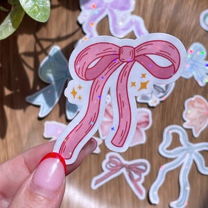 May include: A collection of bow stickers in various pastel colors, including pink, blue, and purple. The stickers have a holographic glitter effect and are scattered on a wooden surface. The central sticker is a pink bow with a white outline and star accents.