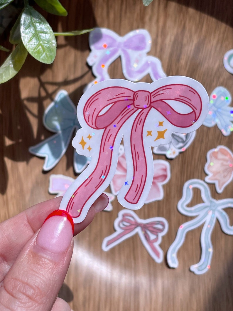 May include: A collection of bow stickers in various pastel colors, including pink, blue, and purple. The stickers have a holographic glitter effect and are scattered on a wooden surface. The central sticker is a pink bow with a white outline and star accents.