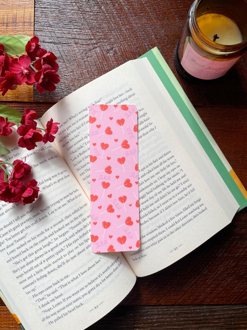 May include: A pink bookmark with red heart and swirl designs is in an open book. Red flowers and a lit candle are on a wooden surface. The bookmark is a reading accessory, ideal for marking pages.