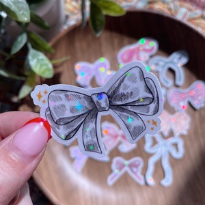 May include: A sticker featuring a detailed black and white bow with a leopard print design. The bow is held in front of a wooden bowl with other bow stickers in various colors and holographic accents. The sticker has star accents.