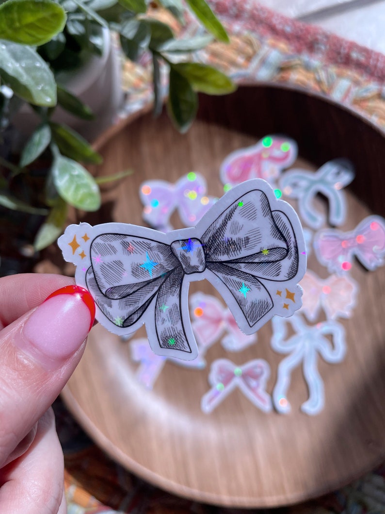May include: A sticker featuring a detailed black and white bow with a leopard print design. The bow is held in front of a wooden bowl with other bow stickers in various colors and holographic accents. The sticker has star accents.