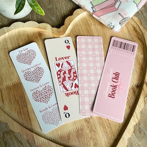 Peut inclure: Quatre marque-pages roses et rouges avec des motifs diff&eacute;rents. Un marque-page pr&eacute;sente un c&oelig;ur avec le texte "Age Gap", un autre marque-page pr&eacute;sente un c&oelig;ur avec le texte "Grumpy Sunshine", un autre marque-page pr&eacute;sente un c&oelig;ur avec le texte "Enemies To Lovers", et un autre marque-page pr&eacute;sente un c&oelig;ur avec le texte "Friends To Lovers". Le quatri&egrave;me marque-page pr&eacute;sente une carte de jeu de la reine de c&oelig;ur avec le texte "Lover" et "Book". Le dernier marque-page est rose avec du vichy blanc et le texte "Romance Reader Book Club".