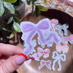 May include: A light purple bow sticker with glitter accents is held in front of a wooden bowl filled with other bow stickers. The bow has a watercolor effect and star details. Other stickers in the bowl are various colors and designs.