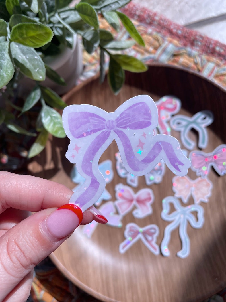 May include: A light purple bow sticker with glitter accents is held in front of a wooden bowl filled with other bow stickers. The bow has a watercolor effect and star details. Other stickers in the bowl are various colors and designs.