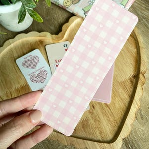 Peut inclure: Un marque-page en gingham rose et blanc avec de petits c&oelig;urs blancs. Le marque-page est fait de carton &eacute;pais et a une surface l&eacute;g&egrave;rement textur&eacute;e.