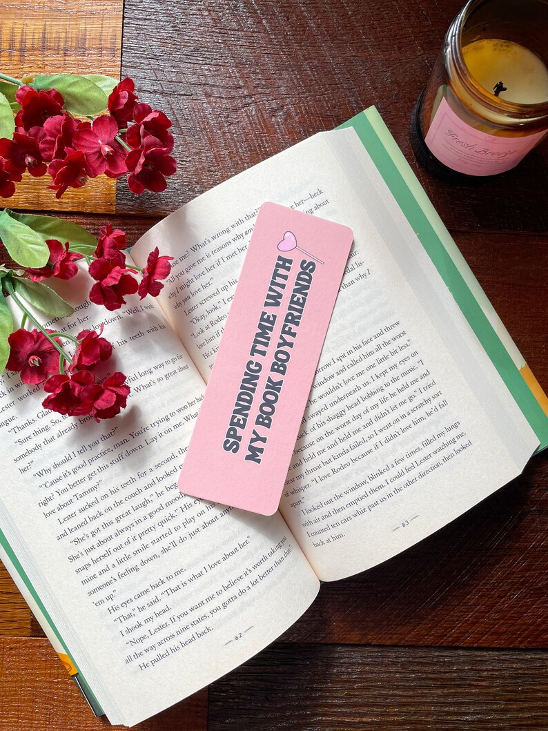 May include: A pink bookmark with the text "SPENDING TIME WITH MY BOOK BOYFRIENDS" and a heart design, placed on an open book. Red flowers and a candle are visible in the background.