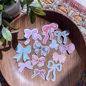May include: A collection of bow-shaped stickers in various pastel colors, including pink, blue, and lavender, are arranged on a wooden plate. Some bows have glitter accents. The stickers are a craft or stationery item.