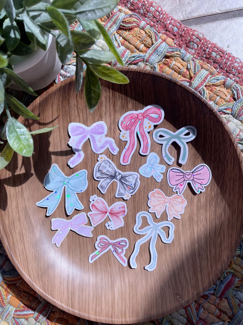 May include: A collection of bow-shaped stickers in various pastel colors, including pink, blue, and lavender, are arranged on a wooden plate. Some bows have glitter accents. The stickers are a craft or stationery item.