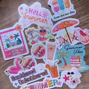 May include: A collection of colorful summer-themed stickers with holographic glitter accents. Designs include phrases like "Hello Summer," images of popsicles, a flamingo, and beach scenes. The stickers are on a wooden surface.