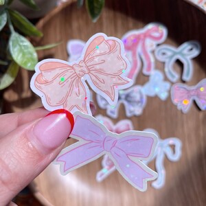 May include: A collection of pastel-colored bow stickers with glitter accents. The stickers feature various shades of pink, peach, and iridescent finishes. Some stickers are held by a hand, while others are scattered on a wooden surface.