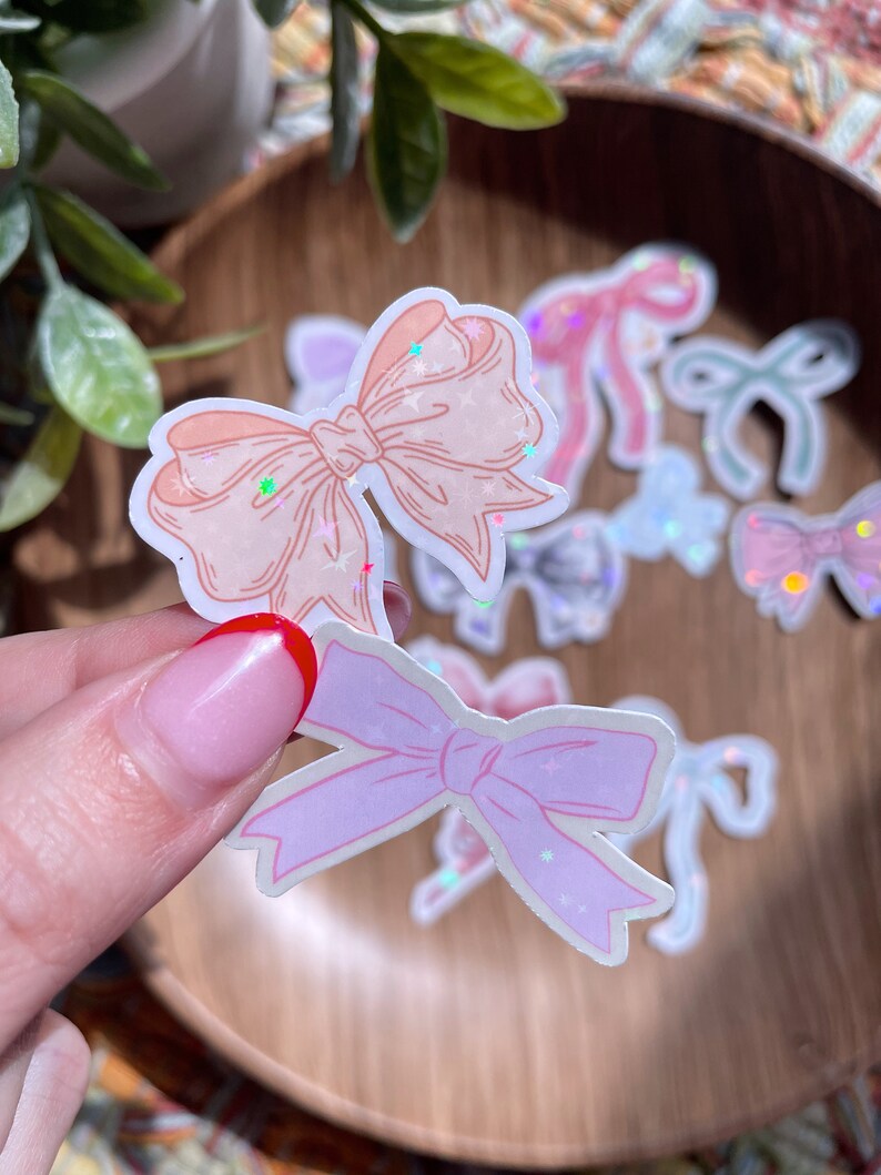 May include: A collection of pastel-colored bow stickers with glitter accents. The stickers feature various shades of pink, peach, and iridescent finishes. Some stickers are held by a hand, while others are scattered on a wooden surface.