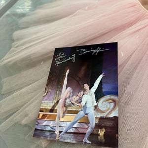 May include: A rectangular photograph featuring a ballet performance with two dancers on stage, signed by Lori Hernandez, resting on a pale pink tulle tutu. The dancers are in costume, with one in a pink tutu and the other in a white and blue outfit.