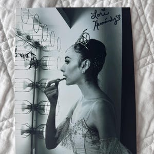 May include: Black and white photograph of a ballerina applying lipstick. She wears a jeweled tiara, a detailed bodice, and a tulle skirt. The photo is signed "Lori Hernandez".
