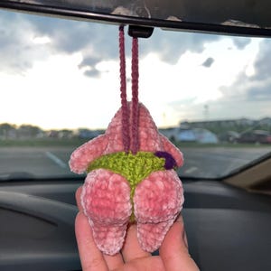 May include: A pink, plush, crocheted car air freshener shaped like a cartoon character's posterior. The character is wearing green shorts with a purple accent. The air freshener hangs from a maroon string.