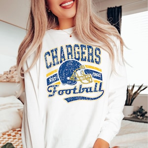 May include: A white long-sleeve shirt featuring a distressed blue and yellow graphic print. The design includes the text CHARGERS, EST 1960, and Football, centered around a blue football helmet illustration with yellow accents and a yellow facemask.