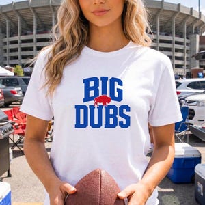 May include: A white short-sleeved t-shirt featuring the text BIG DUBS in bold blue block letters. Between the words BIG and DUBS is a small red silhouette of a buffalo. The shirt is made of a plain fabric and has a standard crew neckline.