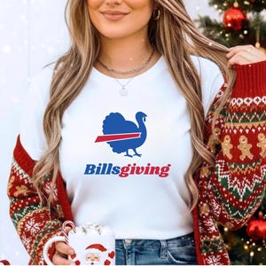 May include: A white short-sleeved t-shirt featuring a blue turkey graphic with a red horizontal stripe across its body. Below the turkey, the text "Billsgiving" is printed in blue and red lettering. The shirt is worn under a red patterned cardigan.