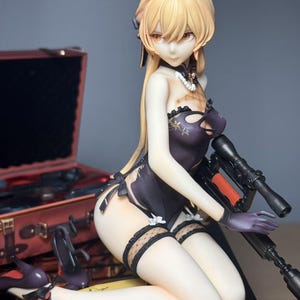 Peut inclure: Figurine détaillée d'une femme aux cheveux blonds, vêtue d'une tenue violet foncé et tenant un fusil noir. La figurine est agenouillée sur une surface noire, avec une valise rouge et des talons hauts noirs en arrière-plan.