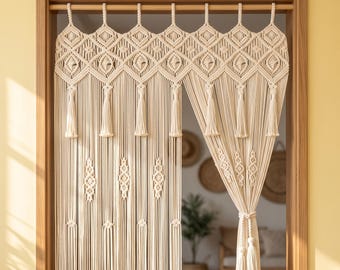Puede incluir: Una cortina de puerta de macramé color hueso con intrincados patrones de nudos, un borde superior en forma de diamante y borlas colgantes. La cortina cuelga de una barra de madera en una puerta, con un lado recogido y atado con un cordón.
