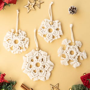 May include: Four handmade macrame snowflake ornaments with wooden beads and hanging cords. The off-white snowflakes have a fringed edge and are arranged on a yellow background with other holiday decorations, including wooden stars and a pinecone.