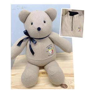 May include: A tan teddy bear with a navy blue ribbon around its neck and a patch on its chest. The bear has a black nose and button eyes. A tan quarter-zip fleece with a logo is in the upper right corner.
