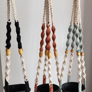 May include: Three macrame plant hangers with wooden beads. The hangers are made with natural cotton rope and feature different coloured accents: black, orange, and green. Each hanger is holding a black plastic pot.